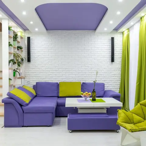 A living room with a purple sectional sofa, lime-green curtains and accent chair, a white painted brick wall