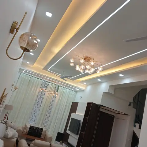 A living room with a stepped cove-lit ceiling, a gold multi-globe chandelier, and a dark wood-veneer TV unit and wardrobe wall, shot at a sharp upward tilt.