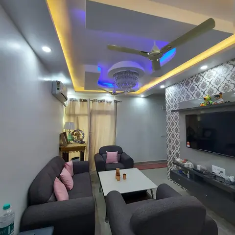 A living room with a stepped POP false ceiling lit in warm and blue LED cove lighting, a crystal chandelier, grey sofas with pink cushions