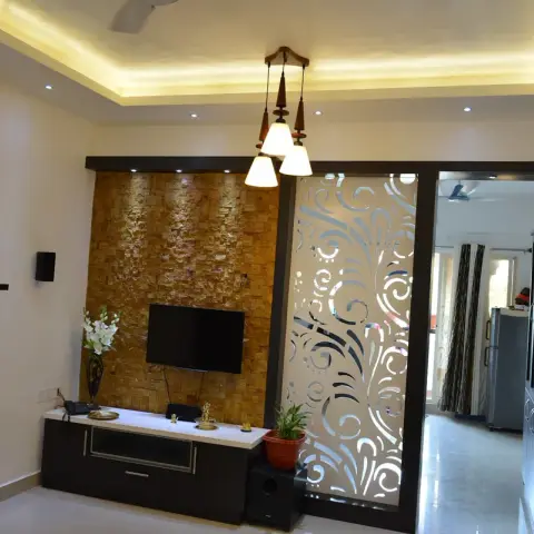 A living room with a stone-clad TV feature wall, a dark wood TV console, and a decorative floral-cutout jaali sliding door leading to the next room