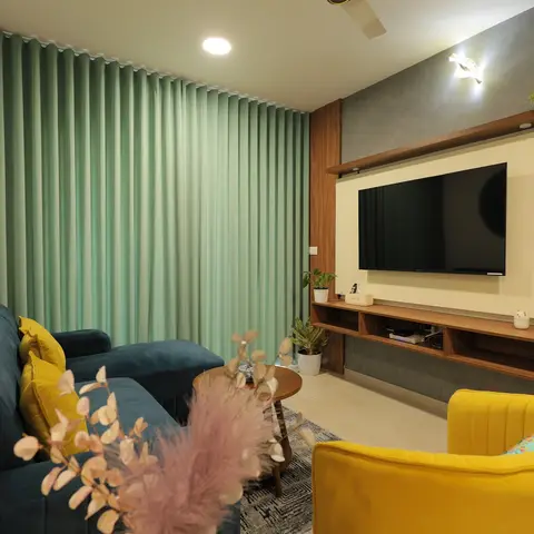 A living room with a teal velvet sectional and mustard armchair facing a floating walnut TV console, sage curtains and a round mandala wall plate