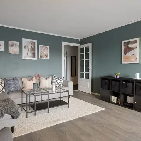 A living room with a teal-grey accent wall, a grey sectional sofa with a sheepskin throw, nesting glass coffee tables, and a dark cube storage console.