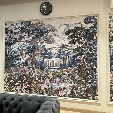 A living room with a three-panel framed scenic garden mural wallpaper spanning the wall, above a tufted charcoal-grey chesterfield-style sofa.