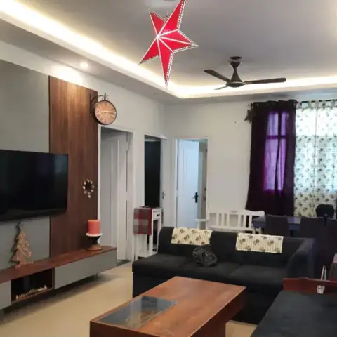 A living room with a vertical fluted-wood TV feature wall, cove-lit ceiling, charcoal sectional sofa and purple curtains, dining table visible beyond.