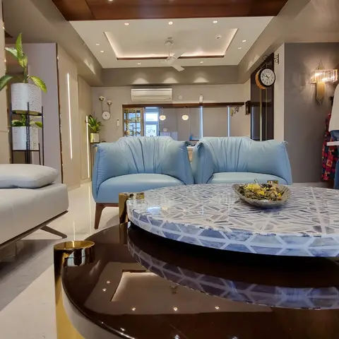 A living room with a wood-clad cove-lit ceiling, powder-blue barrel-back armchairs, and a round blue-and-white mosaic coffee table on brass cylinder legs.