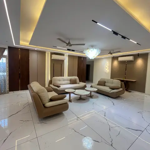 A living room with an angular cove-lit false ceiling, a scalloped glass chandelier, brown leather sofas around round marble-top coffee tables