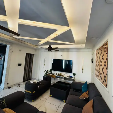A living room with an angular cove-lit false ceiling above a charcoal L-shaped sofa with gold-tipped legs, a black coffee table on marble flooring