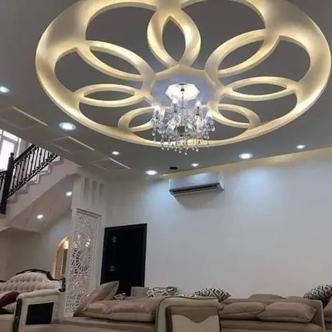 A living room with an elaborate flower-shaped POP false ceiling in cove lighting centered on a crystal chandelier