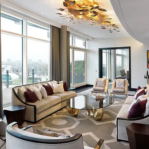 A living room with floor-to-ceiling city-view windows, a sculptural gold pendant chandelier, cream sofas with burgundy accent cushions