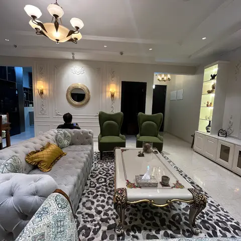 A living room with ornate raised-panel wall moulding and a round mirror, olive-green wingback chairs and a tufted sofa around a leopard-print rug and a