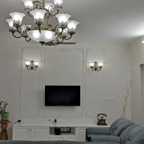 A living room with raised classic panel moulding behind a wall-mounted TV, flanked by ornate brass wall sconces, an ornate crystal chandelier overhead
