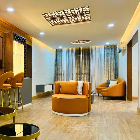 A living room with round orange swivel armchairs, ombre sheer curtains, and a geometric wood-lattice ceiling light panel above a small bar nook with a lit wine
