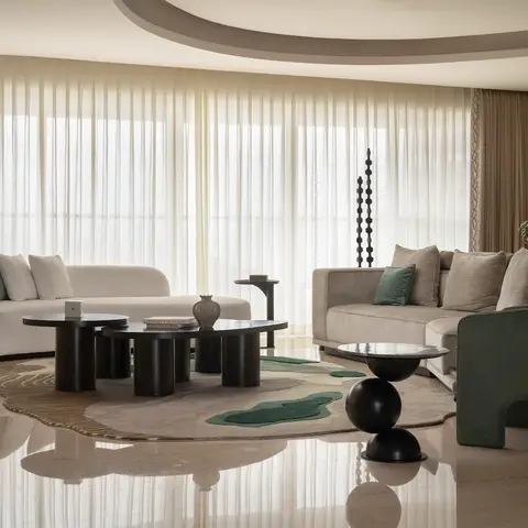 A living room with sculptural black pedestal coffee and side tables on an organic-shaped rug, floor-to-ceiling sheer curtains, and cream and sage-green sofas.