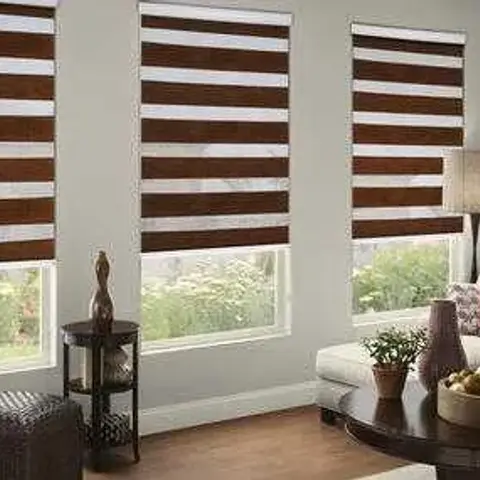 A living room with three matching two-tone zebra roller blinds across the windows, a dark grey sectional sofa, and a round wood coffee table with a fruit bowl.