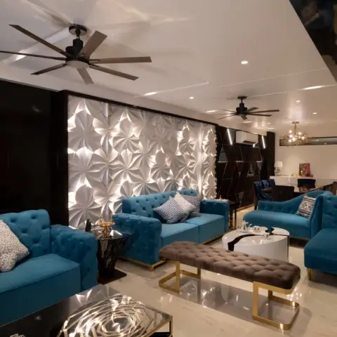 A living room with tufted teal velvet sofas and a gold-framed bench around a round marble coffee table, lit by a backlit 3D origami-pattern feature wall.