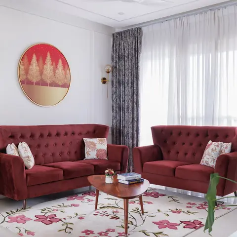 A living room with two maroon tufted sofas facing a wooden coffee table on a pink floral rug, beneath a round gilt-framed print on a wainscoted wall.