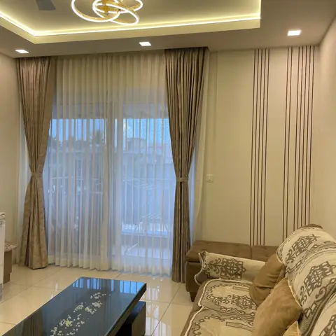 A living room with vertical wood fluted panelling on one wall, a ring-cluster LED pendant hung from a cove-lit ceiling, sheer and heavy curtains