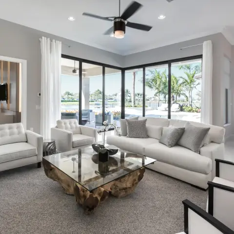 A living room with white tufted armchairs and a sofa around a glass-and-driftwood coffee table, floor-to-ceiling windows framing a poolside palm-tree view.