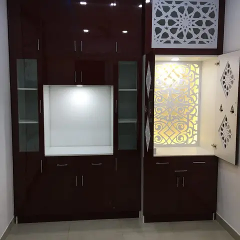 A maroon high-gloss pooja unit with a CNC-cut geometric jaali screen above and a backlit mandir niche with hanging bells behind an open white fretwork door