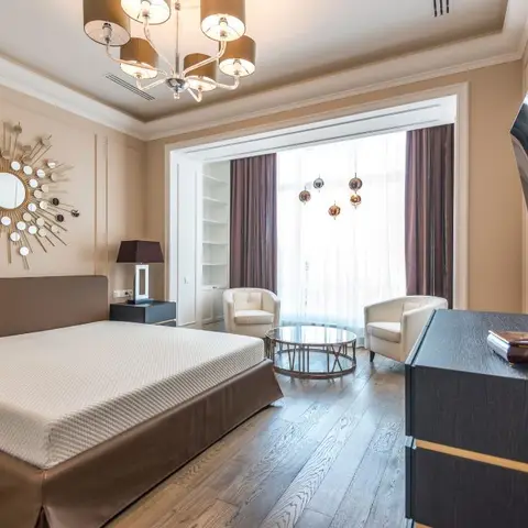 A master bedroom with an upholstered brown bed beneath a sunburst mirror, and an adjoining sitting nook with cream armchairs and a glass coffee table by a