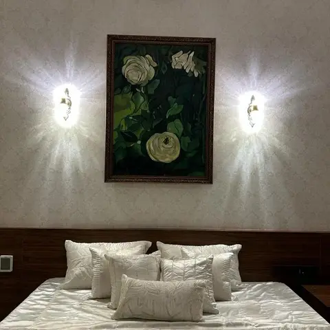 A master bedroom with damask wallpaper, a wood-panelled headboard, symmetrical wall sconces flanking a framed floral painting, and layered white pillows