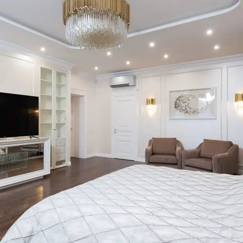A master bedroom with quilted white bedding, a tiered crystal chandelier, moulded wall panelling with brass wall sconces flanking framed art