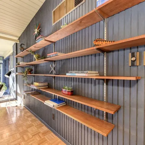 A mid-century open wall-mounted teak shelving unit with brass wall sconces against dark grey slatted panelling, styled with books and a small potted plant