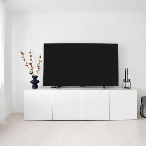A minimalist living room corner with a black flat-screen TV on a white modular console, sheer curtains, and a single styled vase with dried branches.
