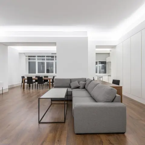 A minimalist open-plan living room with a grey L-shaped sectional sofa, cove-lit ceiling, wood flooring