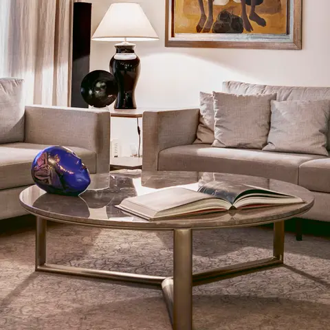 A neutral-toned living room with grey linen sofas around an oval marble-and-brass coffee table styled with a book and a cobalt ceramic mask