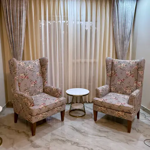 A pair of floral wingback armchairs flank a round brass-and-marble side table in front of layered sheer-and-velvet curtains, on a polished marble floor.