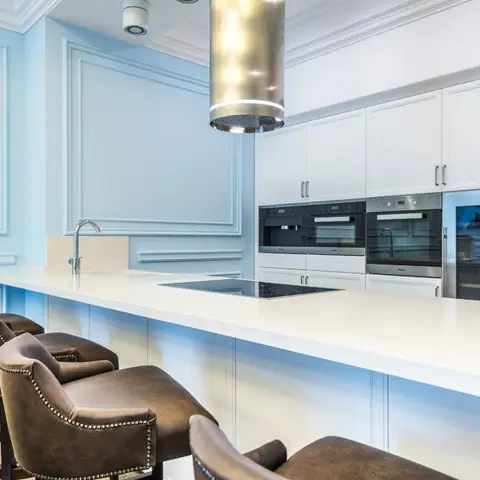 A pastel-blue panelled kitchen island with a white waterfall-edge countertop, integrated wall ovens and a wine fridge, and brown leather nailhead bar stools.