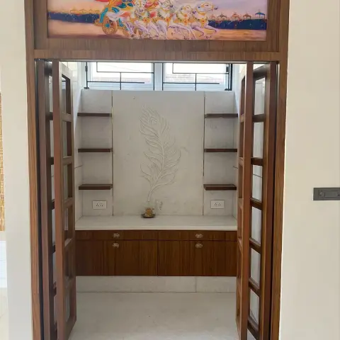 A pooja alcove behind folding glass-and-wood doors, with a hand-carved marble peacock-feather backdrop and a backlit mythological mural above the lintel.