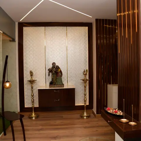 A pooja alcove with a backlit fluted glass panel behind a marble-and-brass idol statue, flanking brass oil lamps, a dark wood-veneer cabinet base