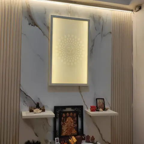 A pooja alcove with a backlit Om motif panel set into a veined marble wall, floating white shelves of deity idols and frames
