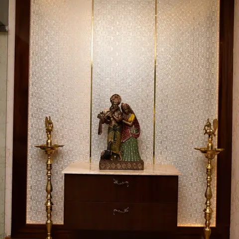 A pooja alcove with a Radha-Krishna brass statue on a marble-topped console, flanked by tall brass diya lamps