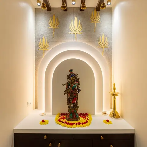 A pooja niche with a backlit arched alcove holding a brass Krishna idol, framed by gold-leaf lotus motifs on a textured stone wall and a beamed wood ceiling