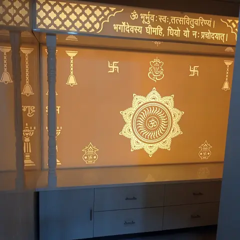 A pooja room with a backlit amber glass panel etched with the Gayatri mantra, an Om mandala and Ganesh motifs above a white storage cabinet