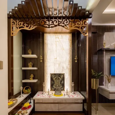 A pooja room with a fan-shaped carved wood arch trimmed in brass filigree, a backlit onyx stone niche