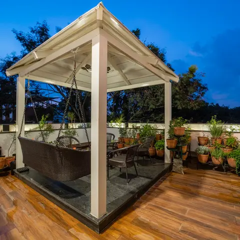 A rooftop terrace at dusk with a white timber gazebo sheltering a hanging rattan swing bed and a rattan dining set, wood-look tile decking