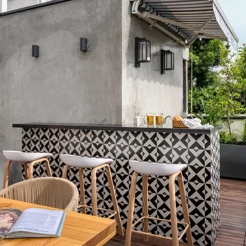 A rooftop terrace bar with a black-and-white geometric tiled counter, bentwood bar stools, a teak dining table