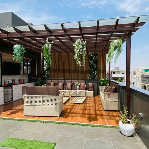 A rooftop terrace lounge with a wood pergola, cream rattan sofas, and cascading potted plants beside a small outdoor bar counter with a mounted TV.