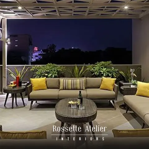 A rooftop terrace lounge with wicker sofas and mustard-yellow cushions beneath a geometric lattice pergola, potted plants lining the parapet at night.