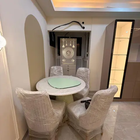 A round marble-top dining table with plastic-wrapped chairs beneath a cove-lit ceiling
