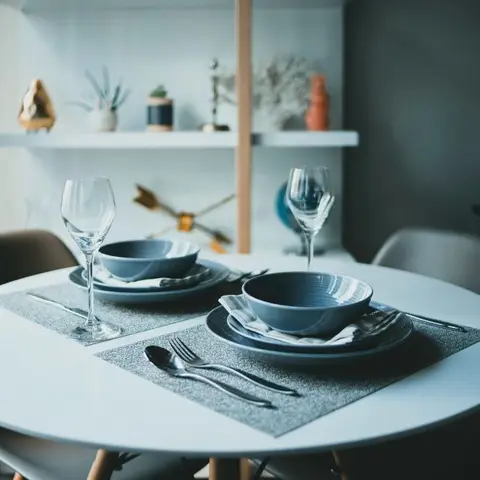 A round white dining table set for two with blue ceramic bowls and wine glasses, wood-legged chairs, styled open shelf behind