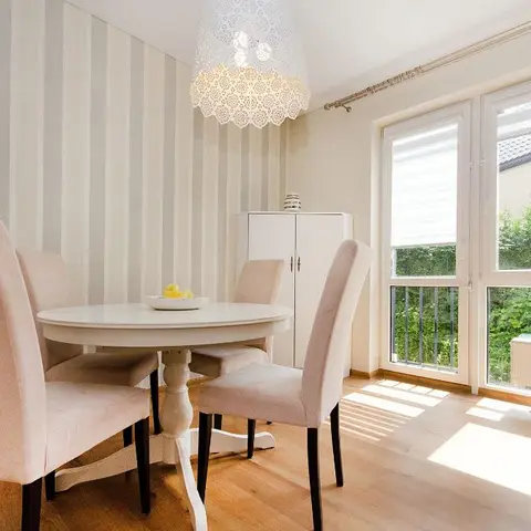 A round white pedestal dining table with four blush-pink upholstered chairs beneath a lace-shade pendant, next to striped wallpaper and a glazed balcony door.