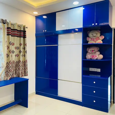A royal-blue and white high-gloss kids' wardrobe wall with open corner display shelves holding teddy bears, beside a matching blue study desk.