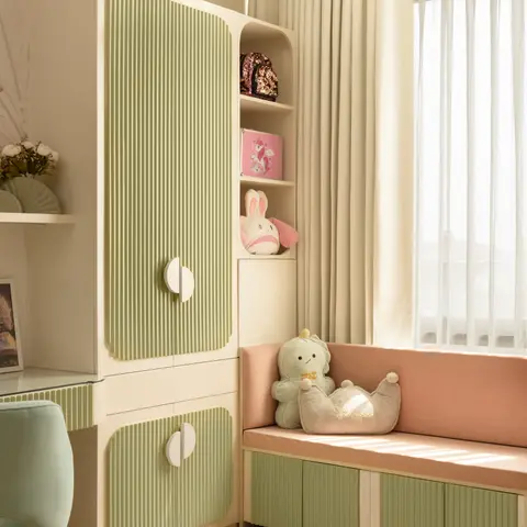 A sage-green fluted-panel wardrobe with a built-in blush window seat and an open toy nook in a girl's bedroom.