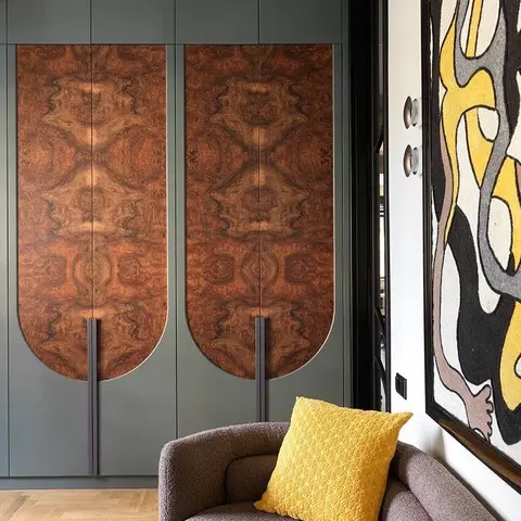 A sage-green joinery wall with arch-shaped burl-wood veneer door panels beside a mauve boucle sofa and a bold abstract textile wall hanging