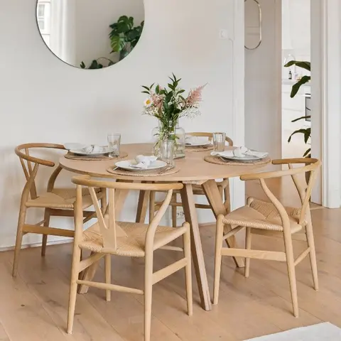 A Scandinavian dining nook with a round light-oak table and woven wishbone chairs under a round wall mirror, set on wide-plank wood flooring.